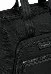 Bolso tote de nylon negro con superficie texturizada, que cuenta con un bolsillo con cremallera y correas verticales. Logo grabado en un parche rectangular.