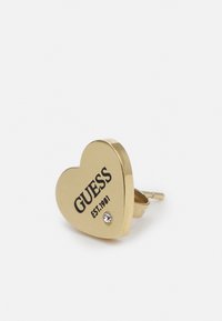 Gouden hartvormige oorbel met zwarte "GUESS" branding en de tekst "EST.1981", voorzien van een kleine heldere steen accent. Gepolijste metalen afwerking.