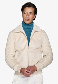 Beige faux fur jakke med en blød tekstur, frontlynlås og en hvid krave. Båret over en lys turkis strikket højhalset sweater.