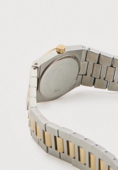 Pulseira de relógio em metal prata e ouro com caixa do relógio fechada, mostrando o verso e uma coroa dourada, sobre fundo branco.