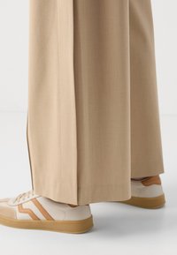 Beige wide-leg trousers with a smooth texture. Paired with white sneakers featuring brown accents and a textured sole.