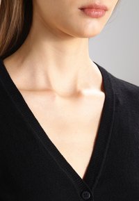 Svart cardigan med V-ringning, ribbad manschett och en jämn textur. Har en enkel knappstängning vid halsen.