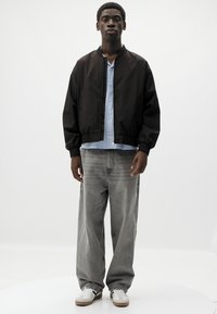 Sort bomberjakke over en lys blå skjorte, parret med grå jeans og hvide sneakers. Jakken har elastiske manchetter og et minimalistisk design.