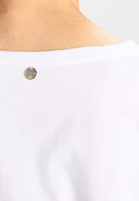 T-shirt bianco in cotone con scollatura rotonda. Un piccolo marchio in metallo è attaccato sul retro, vicino alla scollatura, con dettagli incisi.