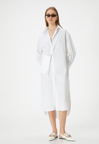 Weißes Hemdkleid mit langen Ärmeln, klassischem Kragen, seitlichen Schlitzen und einer kleinen passenden Umhängetasche. Das Modell trägt weiße Slipper.