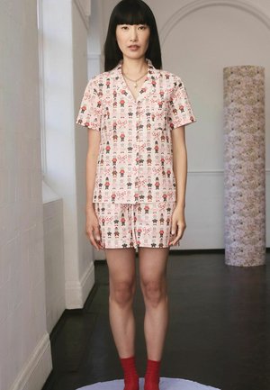 Eine Frau trägt ein passendes kurzärmliges Hemd mit Knopfleiste und Shorts mit einem Cartoon-Charakter-Druck, während sie drinnen auf einer runden lila Plattform steht.