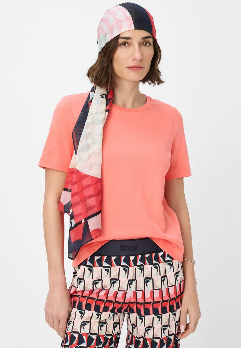 Coral short-sleeve t-shirt paired with patterned trousers featuring geometric designs in red, pink, and navy, complemented by a vibrant patterned scarf.