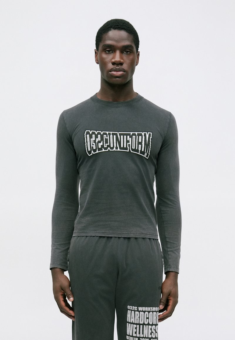 Mand iført en mørk langærmet skjorte med teksten "032C UNIFORM" og matchende bukser med hvid trykt tekst på venstre lår, stående mod en hvid baggrund.