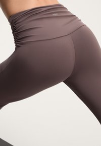 Magas derekú, lila, varrás nélküli leggings, ráncolt elasztikus derékpánttal és sima, rugalmas anyaggal. Minimális márkajelzés látható a deréknál.