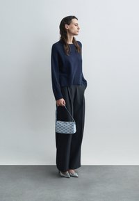 Ναυτικό πλεχτό πουλόβερ, φαρδιά μαύρα παν trousers, ασημένιες γόβες, κρατώντας μια μπλε τσάντα με σχέδιο, που έχει επαναλαμβανόμενο λογότυπο. Απλή φόντο.
