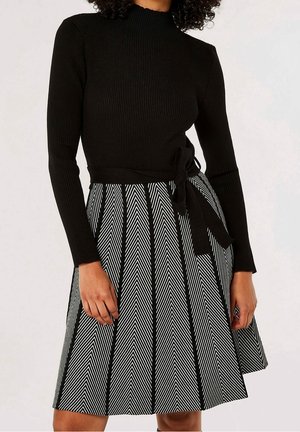 Femme portant un haut à manches longues côtelé noir et une jupe à motif chevron noir et blanc avec une ceinture nouée à la taille.