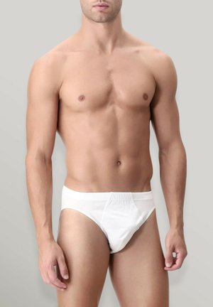 Modello maschile in forma che indossa slip bianchi semplici, in piedi davanti a uno sfondo grigio neutro, con torso e parte superiore delle gambe visibili.