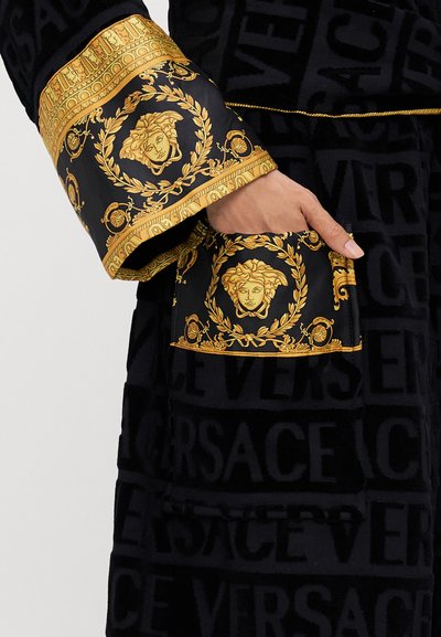 Melna toga ar zeltainiem baroka motīviem, tostarp Medūzas motīviem un tekstu "Versace", ar teksturētu audumu un izteiksmīgiem aproču detalizējumiem.
