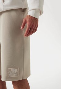 Beige sports shorts made of textured fabric, featuring a patch with logo and year '1981' on the hem, paired with a white long-sleeve shirt.