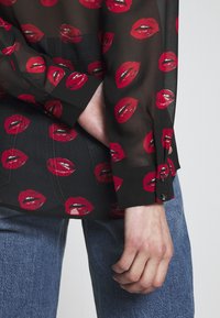 Gennemsigtig sort bluse med røde læbemittryk, lange ærmer med knapdetaljer ved manchetterne, parret med denim. Glat tekstur og letvægtsdesign.