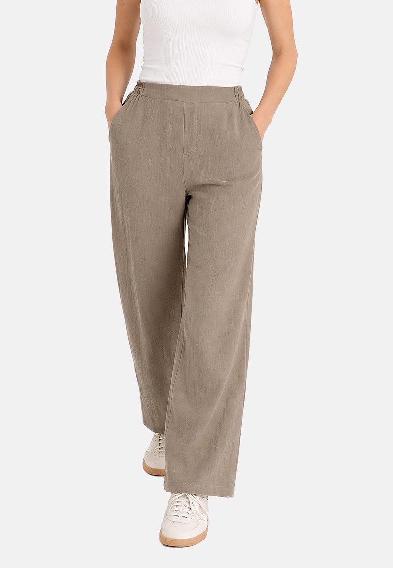 Mujer con pantalones anchos de color marrón claro, camiseta sin mangas blanca y zapatillas blancas, con las manos en los bolsillos, de pie sobre un fondo blanco.