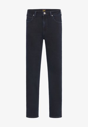 Osoba w oliwkowej koszuli zapinanej na guziki, ciemnych obcisłych dżinsach i czarnych botkach, stojąca na tle jednolitego szarego tła.