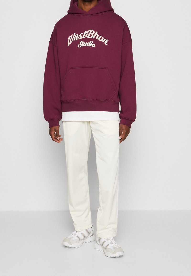 Homme portant un sweat à capuche bordeaux avec poche avant et texte blanc, chemise blanche superposée, pantalon crème et baskets blanches.