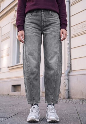 Une personne portant un jean gris ample, un sweat-shirt couleur prune et des baskets blanches se tient sur un trottoir en ville.