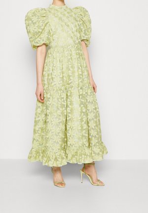 Femme portant une robe à manches bouffantes à fleurs vert clair, avec un ourlet volanté et des sandales à talons assorties, debout sur fond blanc.