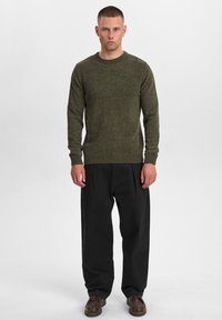 Maglione a maglia verde oliva con scollatura rotonda e finiture a costine, abbinato a pantaloni neri plissettati e scarpe marroni. Tessuto morbido, vestibilità casual.