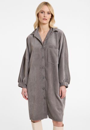 Blonde vrouw die een grijze corduroy blousejurk draagt met lange pofmouwen en een opgestikte zak, gecombineerd met lichtbeige kniehoge laarzen.