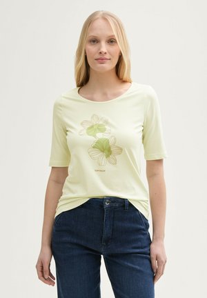 T-shirt jaune clair à manches courtes avec un design graphique floral dans des nuances de vert, associé à un jean en denim foncé.