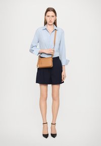 Borsa a tracolla in pelle color tan con chiusura a zip, indossata su una camicia a righe blu e pantaloni corti a pieghe blu navy; presenta una lunga tracolla e dettagli in metallo argentato.