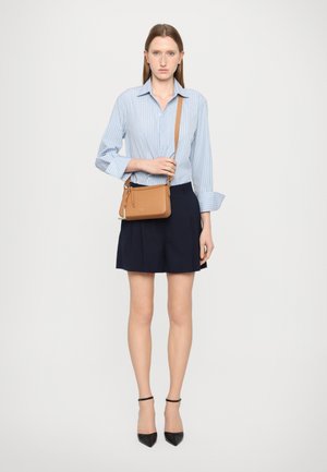 Tan leather crossbody bag with a zip closure, worn on a blue striped shirt and navy pleated shorts; features a long strap and silver hardware.