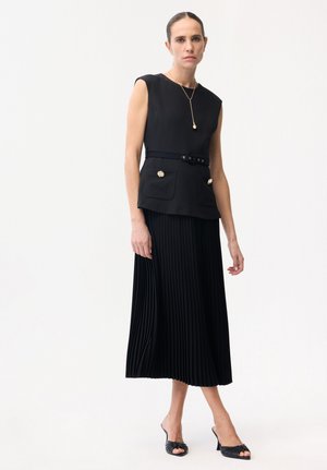 Donna che indossa una maglia senza maniche nera con cintura e bottoni dorati, gonna midi nera plissettata, tacchi aperti neri e una collana con ciondolo dorato.