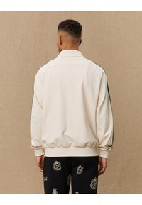 Cream bomber jacket with green side stripes, ribbed cuffs and hem, soft fabric, and a short collar, worn with patterned black pants.
