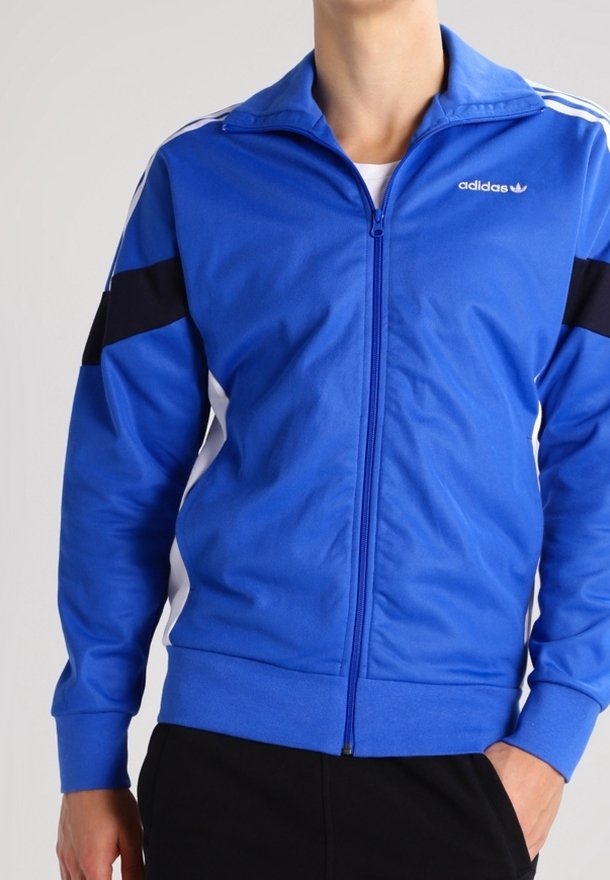 Person som bär en blå Adidas zip-up jacka med vita ränder på axlarna och svarta ränder på ärmarna, tillsammans med svarta byxor.