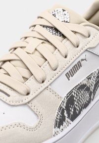 Zbliżenie beżowego i białego sportowego buta Puma z beżowymi sznurówkami i detalem w czarno-białym wzorze wężowym po bokach i na języku.