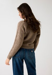 Brązowy sweter z dzianiny z prążkowanym pasem w talii oraz szczegółowym wzorem ściegów wzdłuż rękawów, w połączeniu z niebieskimi dżinsami.