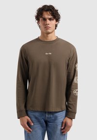 Bruin langärmelig shirt van zachte stof, met een ronde hals en bedrukte accenten op beide mouwen. Gecombineerd met een blauwe spijkerbroek.