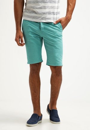 Pantalones cortos de algodón turquesa con un corte holgado, bolsillos laterales y dobladillo ligeramente enrollado, combinados con zapatos oscuros sin cordones y camisa a rayas.