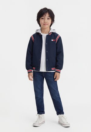 Garçon debout portant une veste varsity bleu marine avec des rayures blanches et rouges, un t-shirt blanc, un jean bleu et des baskets blanches montantes.