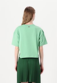 Grønn kortermet bomulls t-skjorte med en cropped design og et merket accent på baksiden. Brukt over et plissert grønt skjørt.