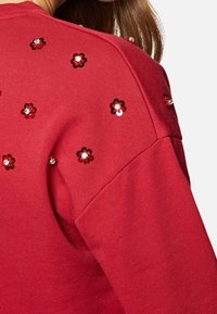 Roter Sweatshirt mit strukturierter Oberfläche, ausgestattet mit dekorativen floralem Pailletten in Schwarz und Rot im Schulterbereich.