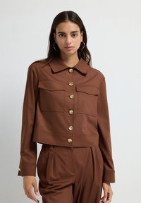 Ung kvinna med långt mörkt hår bär en brun knäppt jacka med stora fickor och matchande högmidjade plisserade byxor.