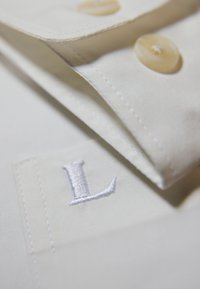 Close-up van een witte shirtkraag met beige knopen en een geborduurde letter "L" nabij de manchet.