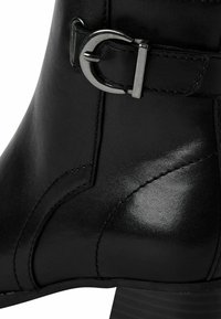 Bota de tobillo de cuero negro con acabado brillante, que presenta un acento de hebilla metálica y costuras detalladas a lo largo del costado. Tacón grueso.
