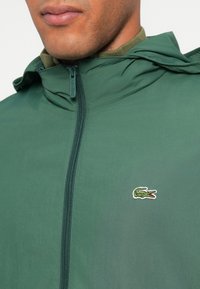 Zelena jakna na patentni zatvarač od laganog materijala; s kapuljačom, zatvaranjem na patentni zatvarač i malim Lacoste krokodilom na prsima.