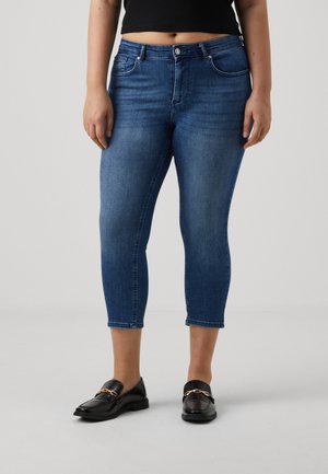 Kvinde iført mørkeblå cropped skinny jeans, sorte loafers med guldkædedetalje og sort cropped top, stående på ensfarvet grå baggrund.