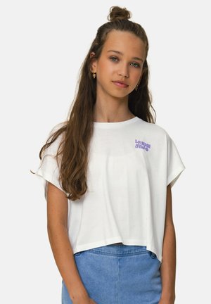 Jonge vrouw met lang haar, gekleed in een wit cropped T-shirt en een blauwe denim rok, staand tegen een effen lichte achtergrond.