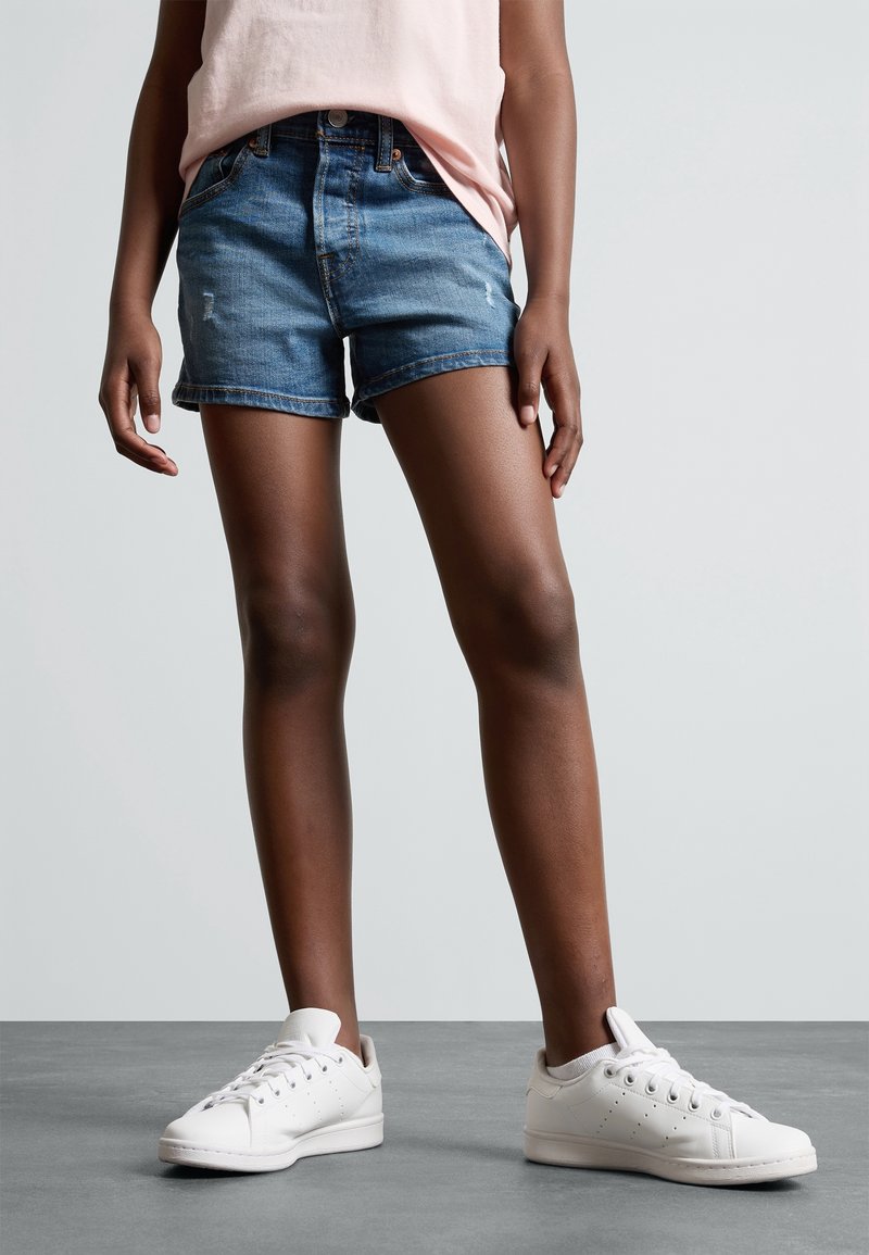 Denimshorts i medelblå med fransad nederkant, i kombination med vita sneakers som har en slät yta och en minimalistisk design.