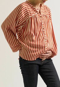 Rood en crèmekleurig gestreept blouse met een V-hals, knoopsluiting en geraffineerde taille, gecombineerd met donkere jeans. Heeft lange, losse mouwen.