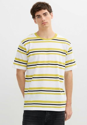 Joven con camiseta blanca de rayas horizontales amarillas y negras, de pie frente a un fondo claro y sencillo.