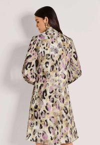 Frau mit langen dunklen Haaren, die goldene Statement-Ohrringe und ein knielanges, leopardfarbiges Kleid in lila und beige Tönen trägt, mit dem Rücken zur Kamera gewandt.