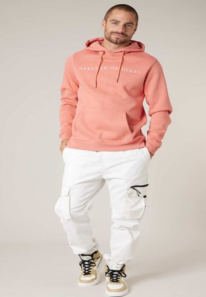 Man draagt een koraalroze hoodie met witte tekst, witte cargobroek met ritssluitingen en beige hoge sneakers, staat met handen in de zakken.